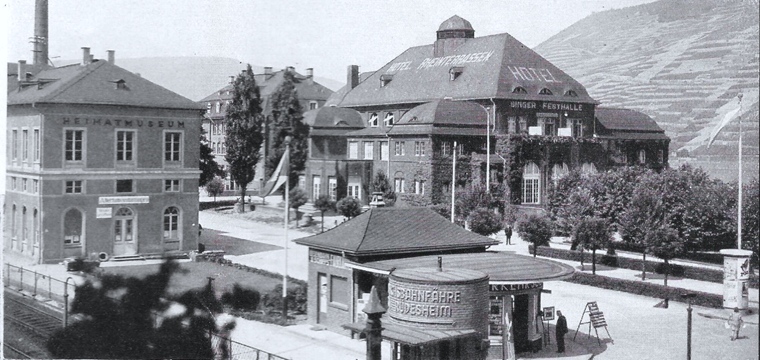 Das Foto aus der Mitte der 1935 Jahre zeigt die Binger Festhalle (Palais Bingen) am Rheinufer und davor, nahe der Gleise, das Badhaus mit dem Heimatmuseum und zuvor der Pavillon mit dem Verkehrsbüro.