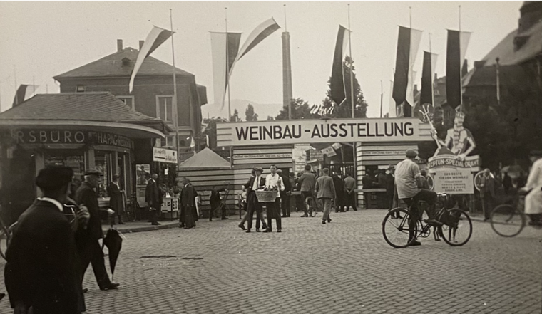 Blick auf den Eingang zur Weinbauausstellung. Links Pavillon und Badhaus, am rechten Rand die Festhalle (später Palazzo, Palais). Der Fotograf steht auf Höhe der Bahnüberführung am Starkenburger Hof, die noch heute existiert.