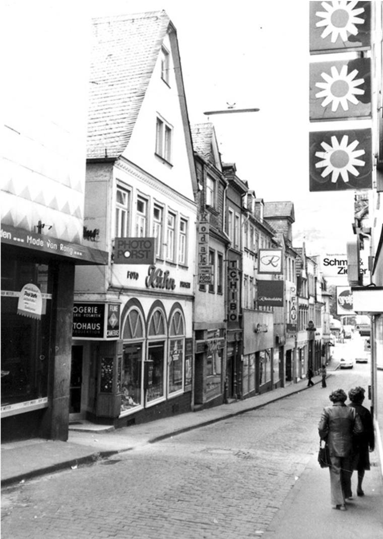 Ca. 1970 mit den bekannten Geschäften Drogerie Kühn, Rheinelektra, Tchibo und Fauck links im Bild. Die Hausecke mit dem Erker gehört zum Herrenhaus des Modehauses Sinn. Das Haupthaus (Damenkonfektion) befand sich gegenüber auf der Südseite des Speisemarkts. Noch mit den schmalen Bürgersteigen und dem alten, unebenen Pflaster, das bei schlechtem oder frostigem Wetter nicht ungefährlich war.