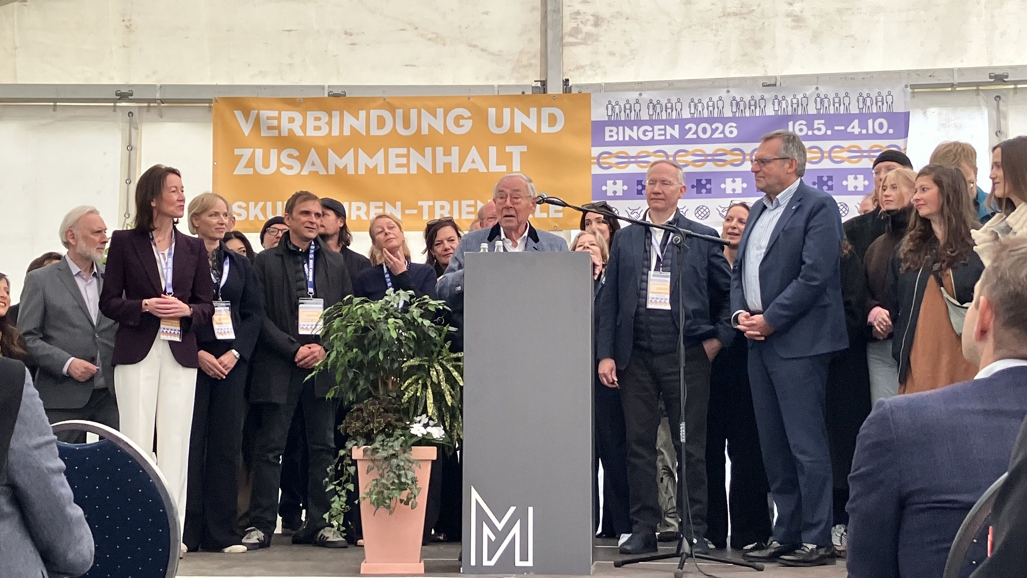 Abschließend stellten sich die Verantwortlichen der Triennale zum Gruppenbild. Mit dabei: Maria Gleichmann-Pieroth (2. v. li.) Kuno Piroth (am Rednerpult) und der Binger Oberbürgermeister Thomas Feser (vorne, blauer Anzug).